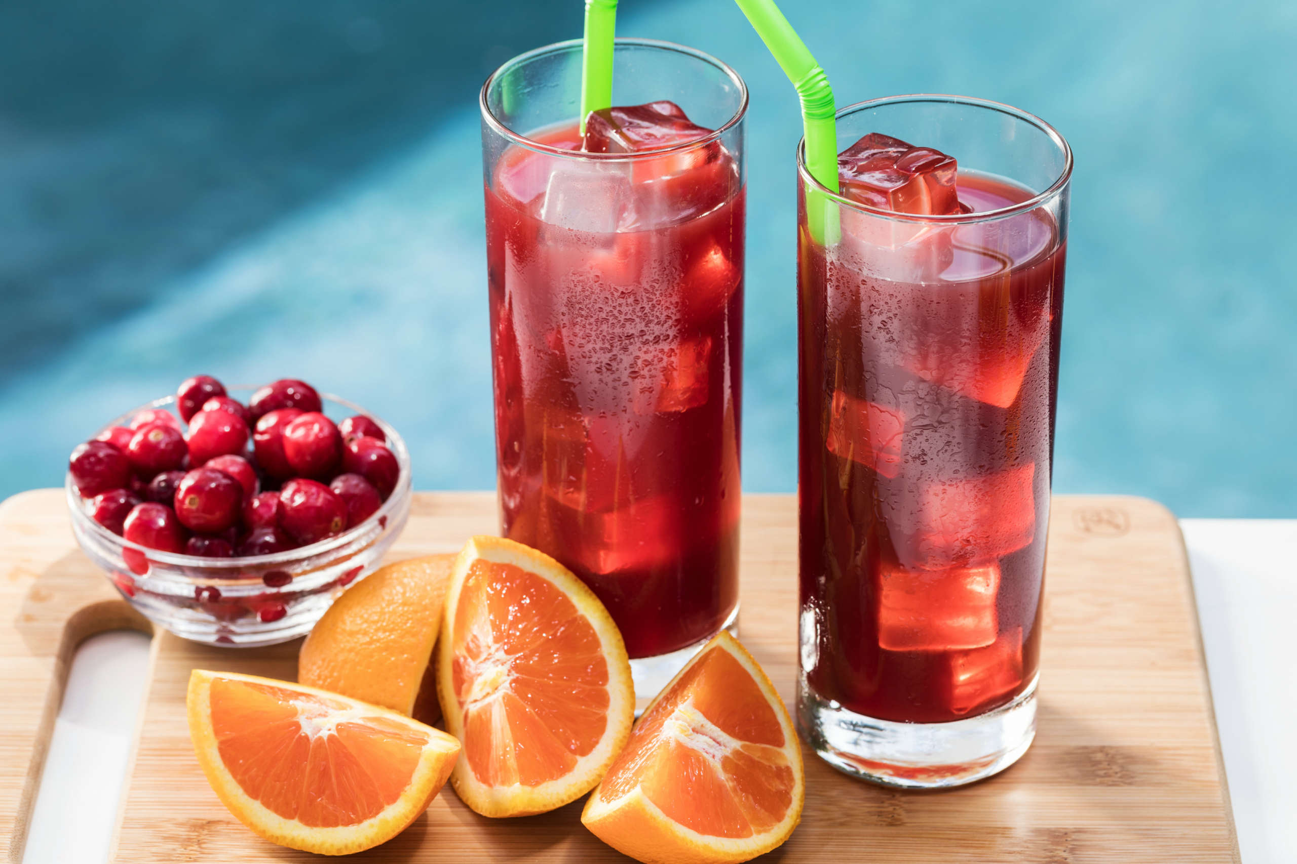 Two tall glasses of bright red Orange Cranberry Iced Tea on ice with straws, next to orange slices