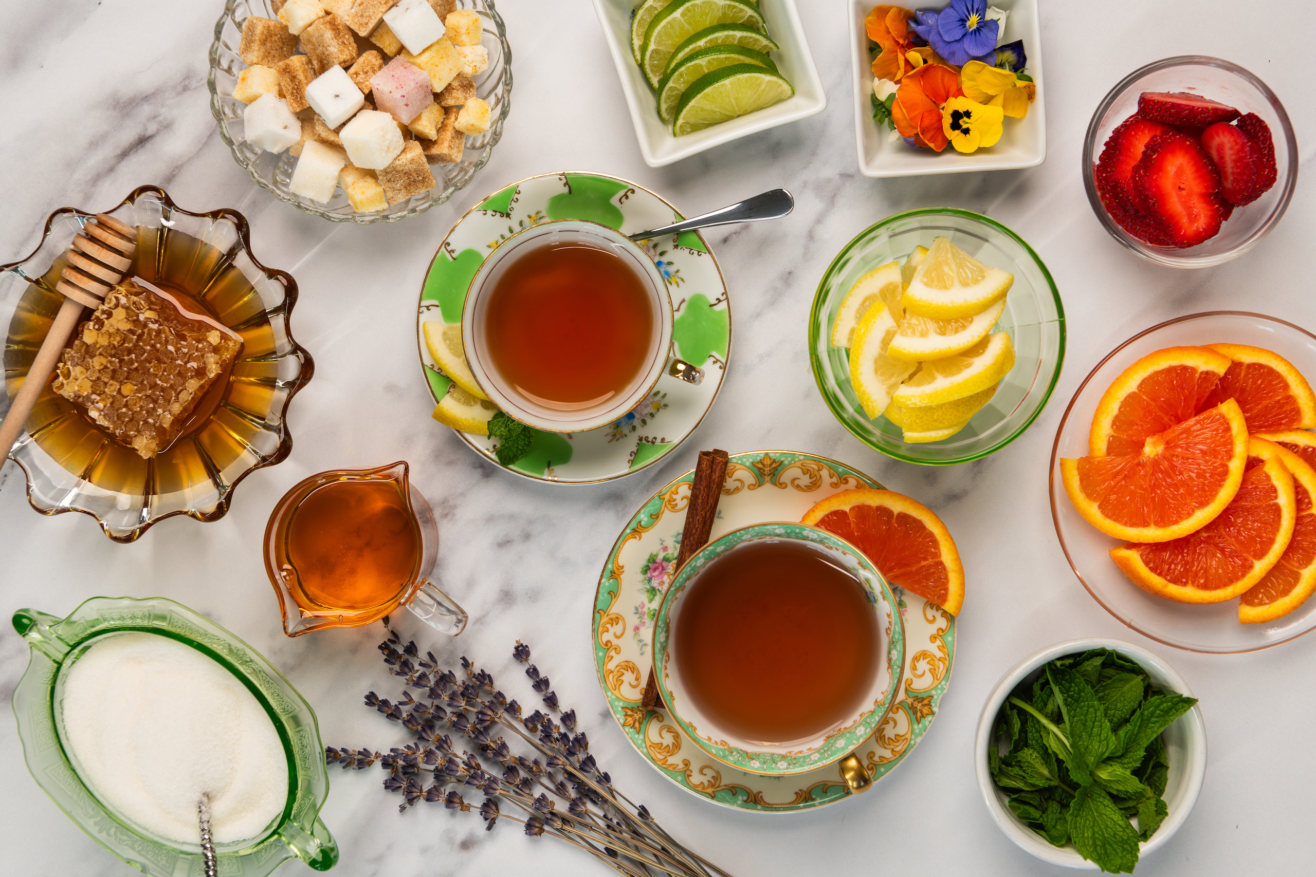 A selection of sweeteners and garnishes laid out for a tea party, including cane sugar, agave syrup, honey and honeycomb, flavor infused sugar cubes, fresh mint leaves, sliced lemon, orange and lime, edible flowers, cinnamon sticks.